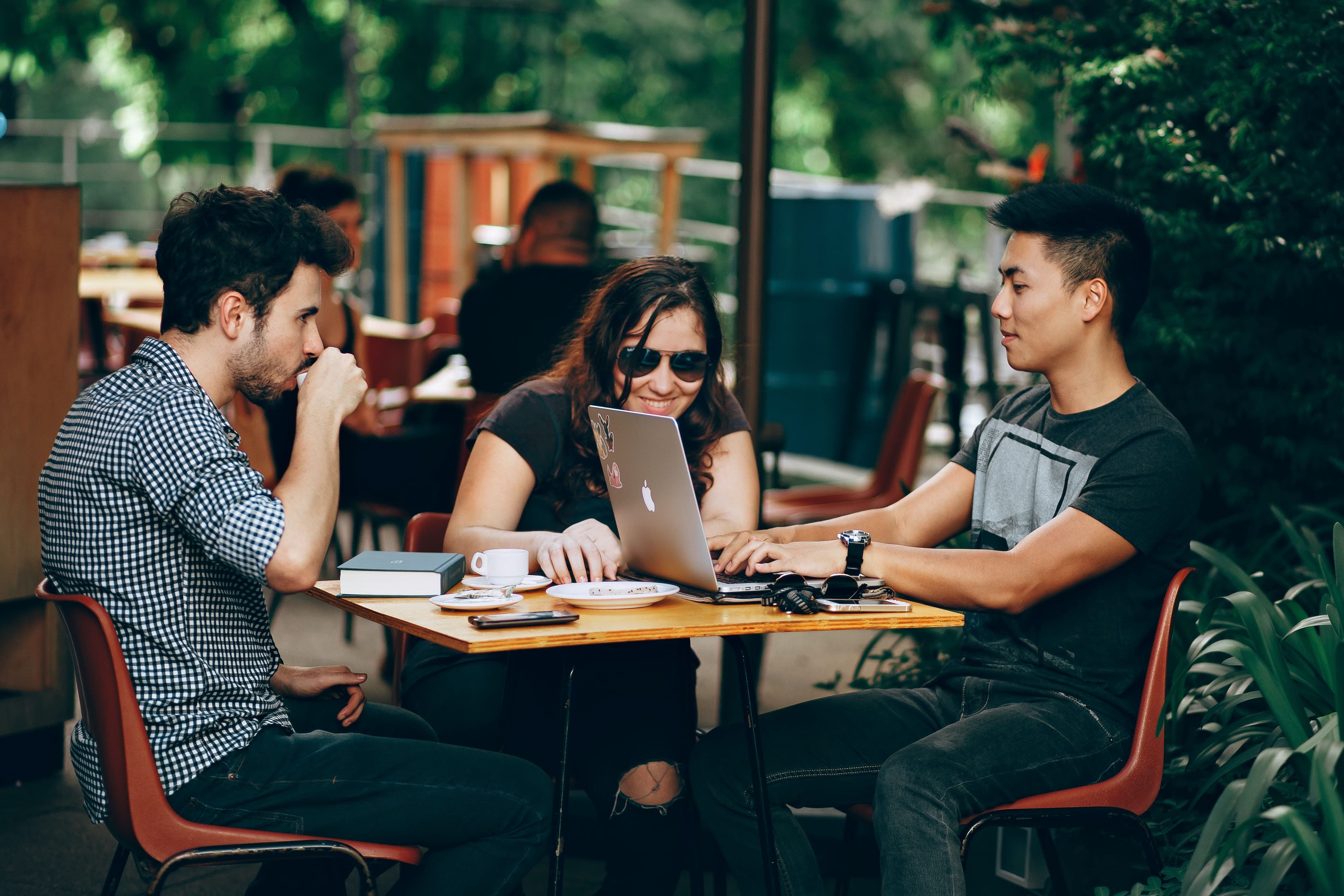 A photo of three colleagues sitting outdoors at a cafe table, engaged in discussion while working on a laptop. The group appears to be collaborating, with a focus on teamwork and problem-solving in a relaxed setting.